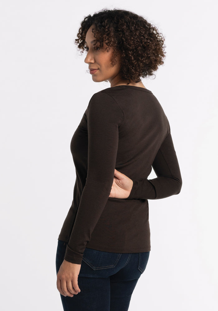 A person with curly hair wearing the Woolx Layla V Neck Top in French Roast and dark jeans stands in a three-quarter view against a plain white background.