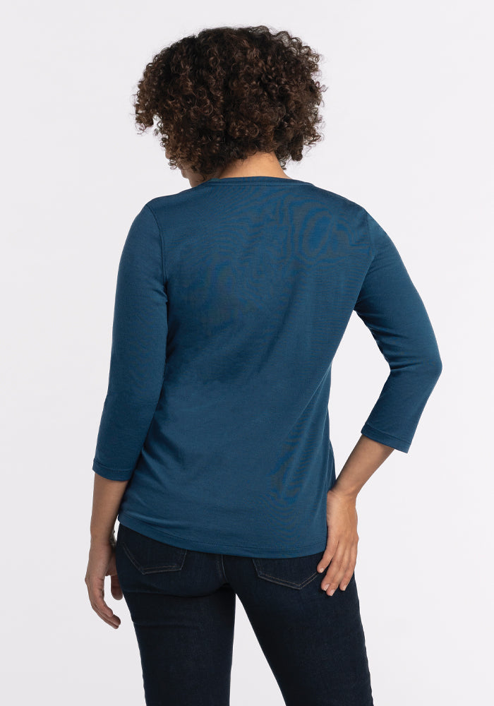 A person with curly hair, dressed in a blue, relaxed-fit Elena V Neck in Real Teal by Woolx and dark pants, stands with their back to the camera against a plain white background.