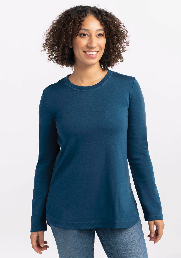 A person with curly hair is smiling in a relaxed-fit, merino wool Hazel Tunic in Real Teal by Woolx, paired with jeans. They are standing against a plain white background. 