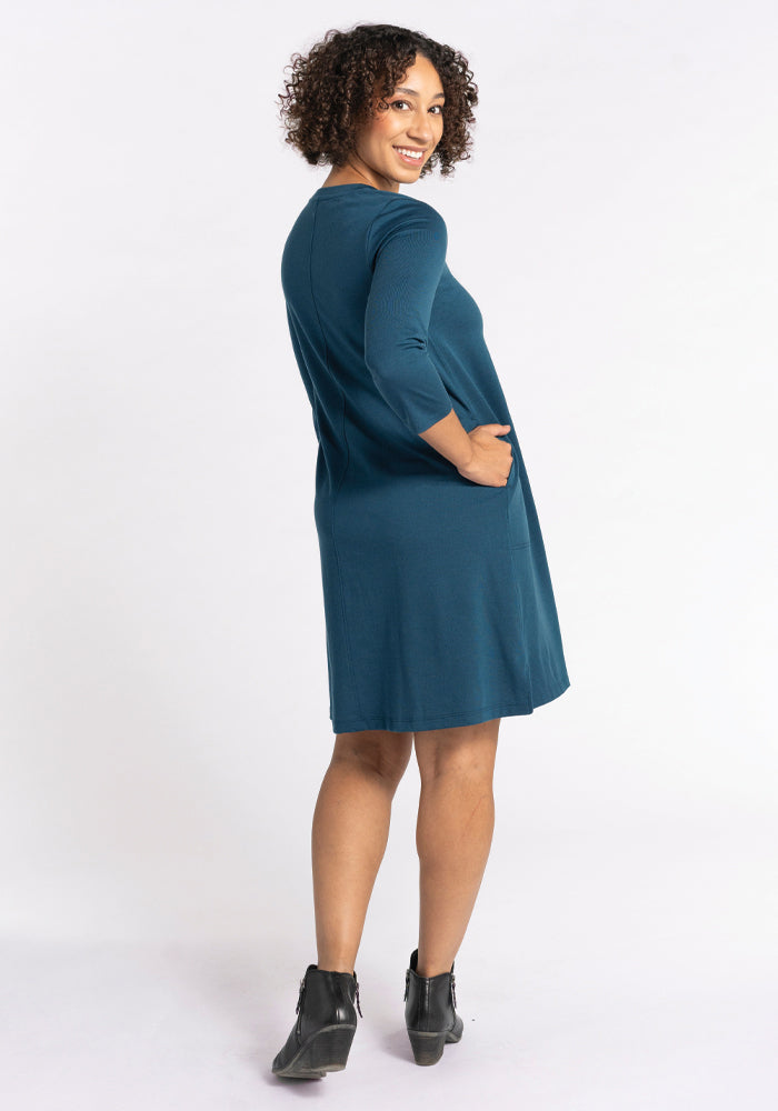 A person with curly hair smiles while wearing the Woolx Rissa A Line Dress in Real Teal, which is perfect for its travel-ready design. With one hand in the dress pocket and standing against a plain white background, they complement the look with ankle boots.