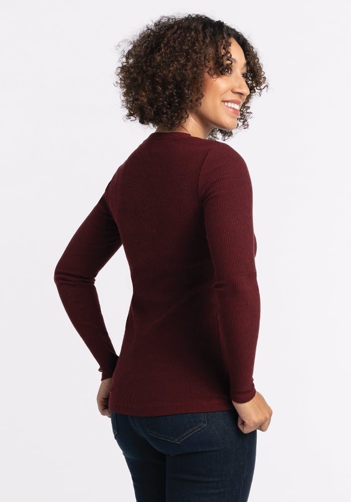 A person with curly hair is smiling and looking to the side, wearing the Hadley Ribbed Crew in Cranberry Melange by Woolx, paired with dark jeans—an ideal choice for a versatile travel wardrobe. The background is plain white.