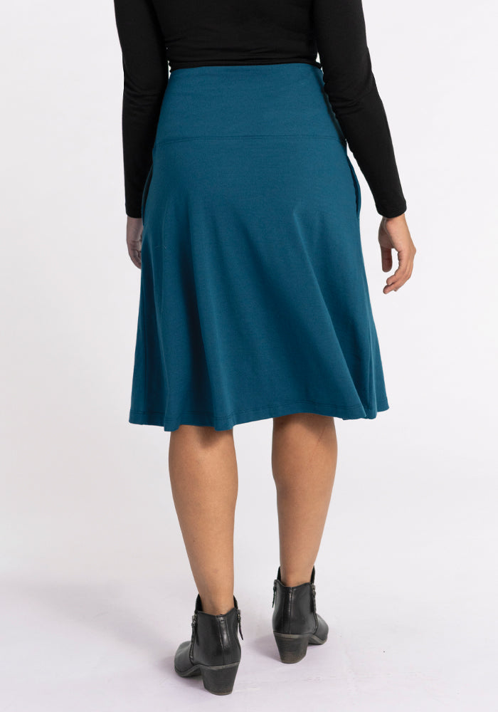 The image shows the back view of a person wearing a Woolx black long-sleeve top paired with the Marilyn Flowy Midi Skirt in Real Teal. They have on black ankle boots featuring wedge heels, against a plain white background.