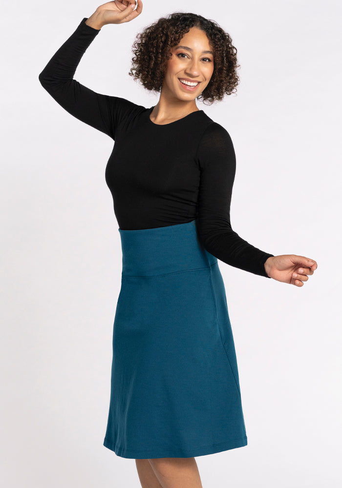 A person with curly hair smiles and poses with one arm raised, showcasing their black long-sleeve top paired with the Marilyn Flowy Midi Skirt in Real Teal from Woolx. The backdrop is a simple white, emphasizing their vibrant outfit choice.