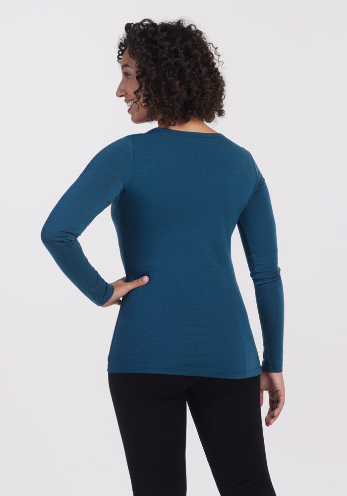 A person with curly hair is wearing the Woolx Layla V Neck Top in Real Teal with black pants, standing with their back to the camera against a plain white background.