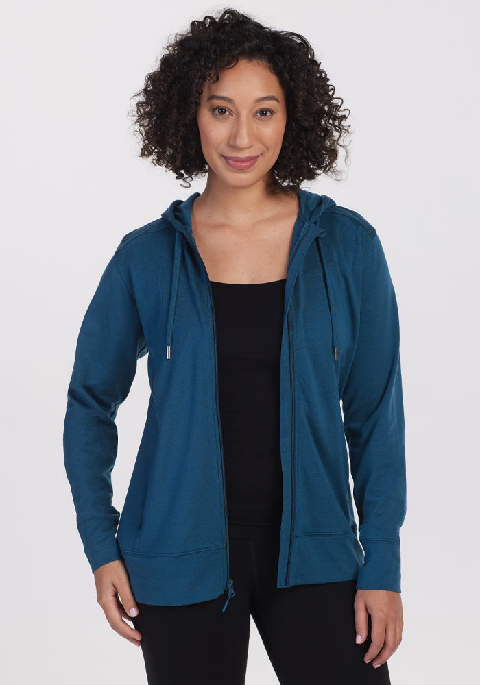 A person with curly hair is smiling at the camera against a plain backdrop, wearing the Ryann Hoodie in its athletic fit and real teal color by Woolx over a black top. 