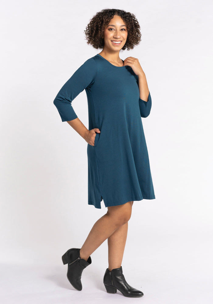 A person with curly hair stands in a Woolx Rissa A Line Dress in Real Teal, crafted from Merino wool. The knee-length dress features three-quarter sleeves and practical pockets, pairing perfectly with black ankle boots. They smile and pose against a plain white background, showcasing a travel-ready style.