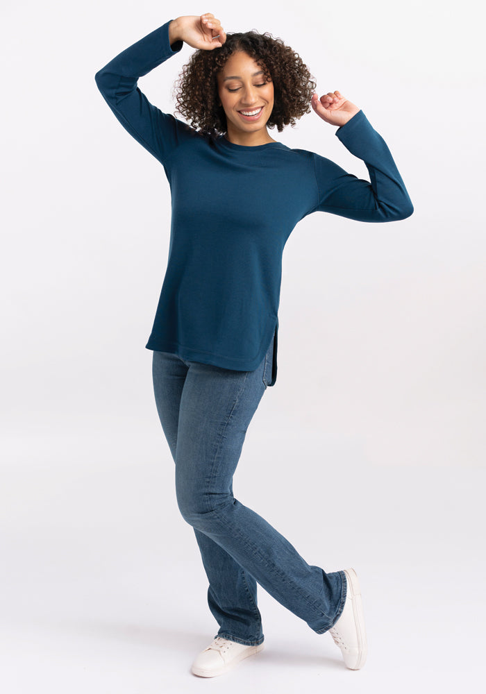 A person wearing the Woolx Hazel Tunic in Real Teal with jeans is smiling and striking a playful pose. They stand on one leg with the other slightly bent, arms raised against a plain white background.