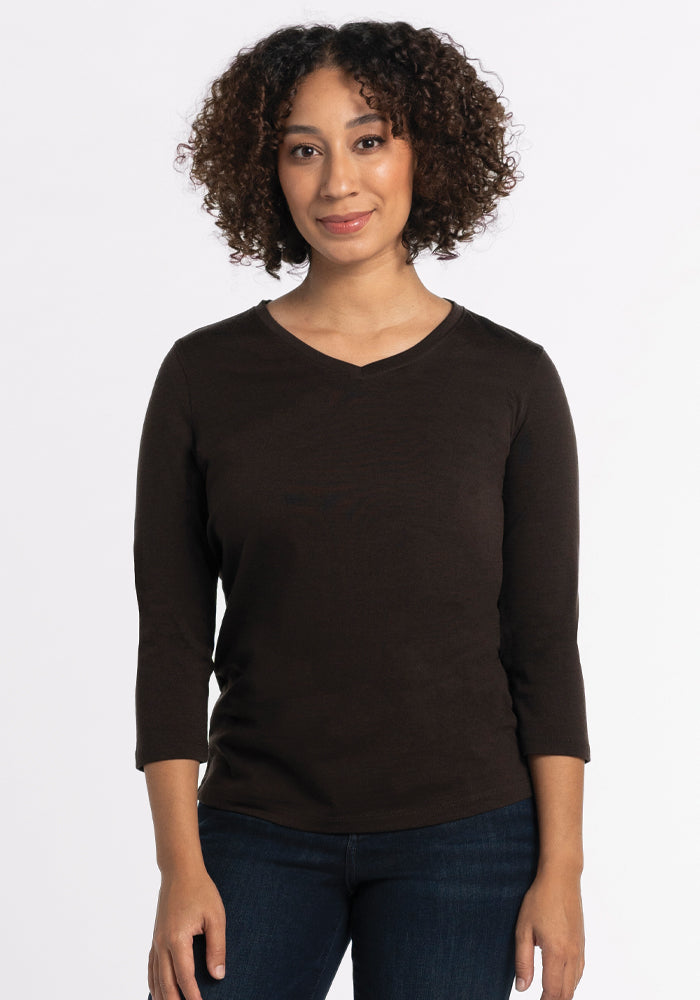 A person with curly hair is wearing a Woolx Elena V Neck in French Roast and dark jeans, standing against a plain white background. 