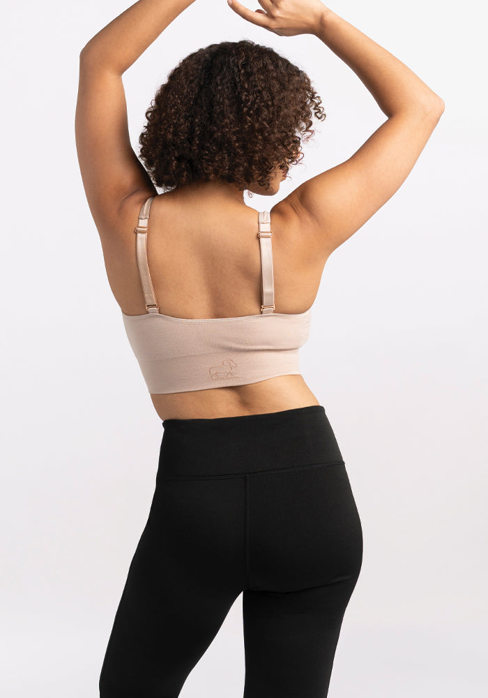 A person with curly hair stands with their back to the camera, arms raised, showcasing a beige sports bra and the flattering bootcut style Marissa Pants in black from Woolx, made from odor-resistant MerinoFlex® fabric, against a plain background.