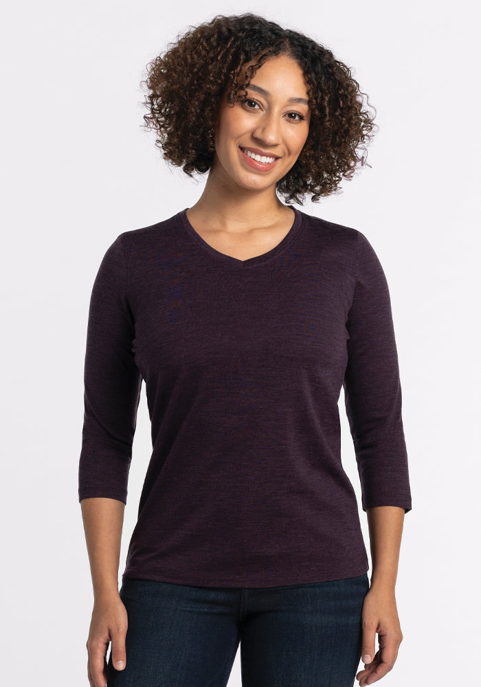 A person with curly hair smiles while wearing a relaxed fit, deep plum Elena V Neck Top by Woolx and dark jeans against a plain white background.