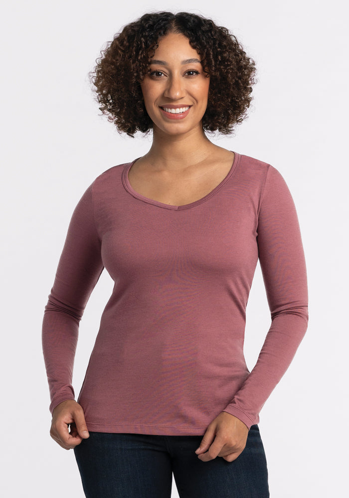 A person with curly hair smiles while standing against a plain white background. They are wearing a Wild Ginger Layla V Neck Top by Woolx and dark jeans, exuding effortless style. 