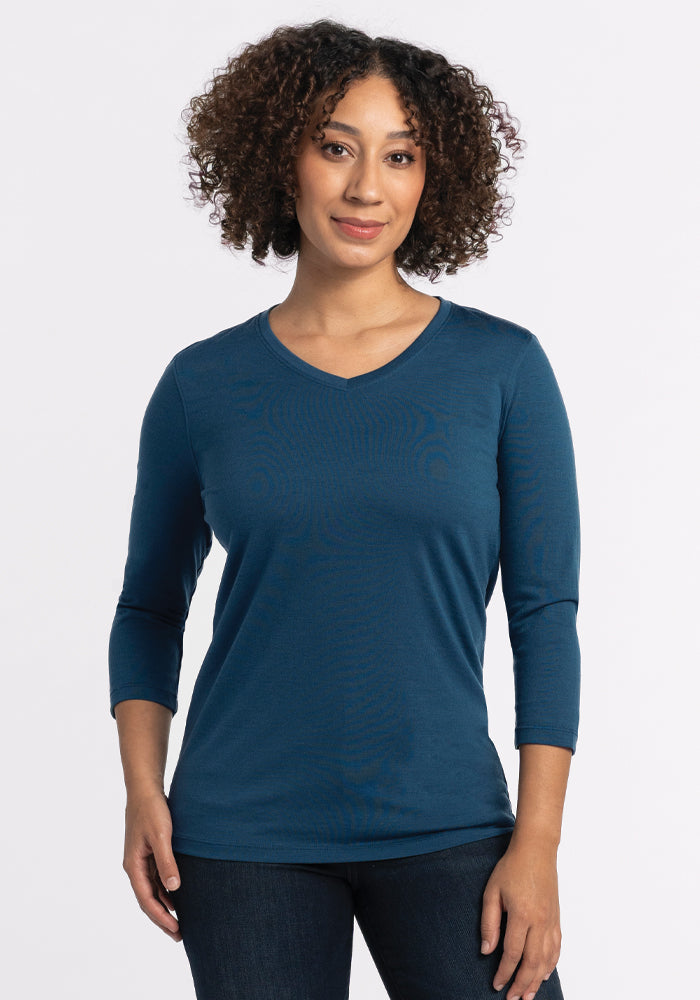 A person with curly hair is wearing a Woolx Elena V Neck in Real Teal, featuring a relaxed fit. They are standing against a plain light background, smiling gently while looking at the camera.