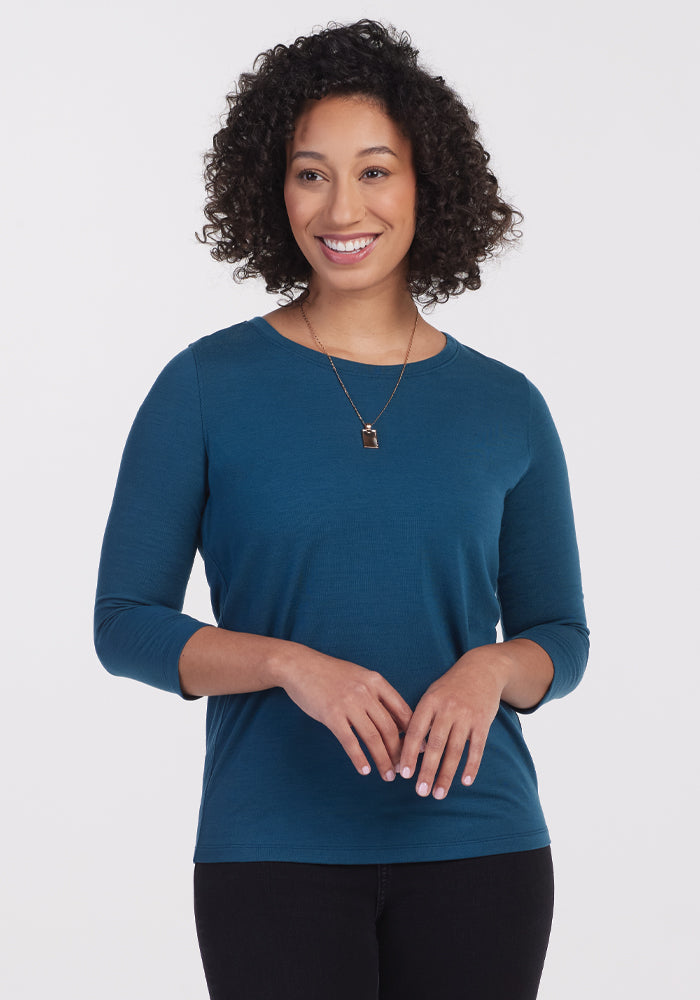 A person with curly hair is smiling and wearing a Jenny 3/4 Sleeve Crew Neck in Real Teal by Woolx, along with a necklace. They stand against a plain white background, highlighting the lightweight shirt's comfort and style.