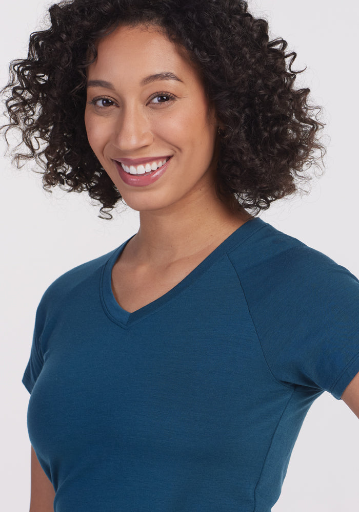 A person with curly hair smiles at the camera, wearing a short-sleeved Mia V Neck in Real Teal from Woolx. The background is plain and white.