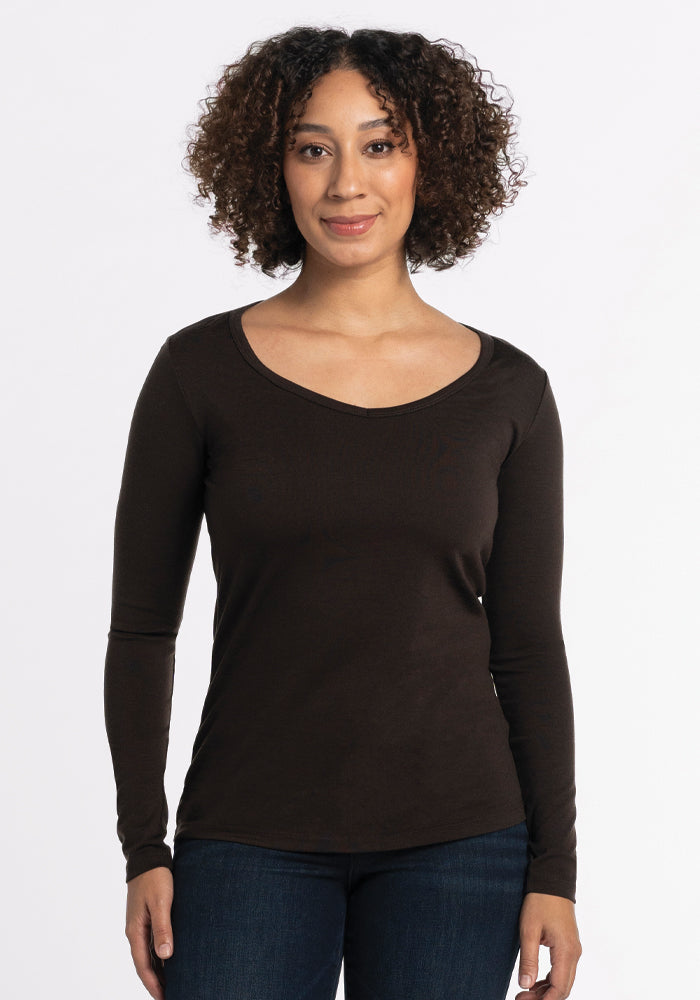 A person with curly hair is wearing the Woolx Layla V Neck Top in French Roast paired with jeans, standing against a plain white background and showcasing the effortless style of a breezy knit.