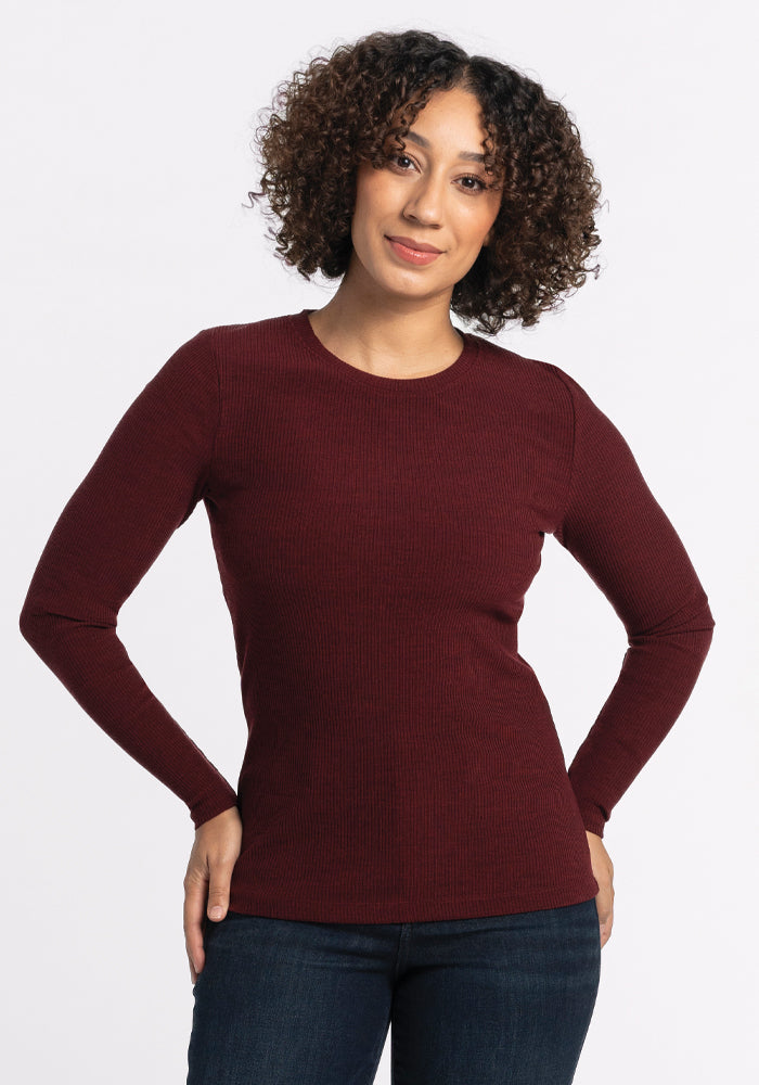 A person with curly hair is wearing the Woolx Hadley Ribbed Crew in Cranberry Melange, paired with dark jeans. They are standing against a plain white background, smiling with hands on hips, perfectly showcasing their stylish travel wardrobe. 