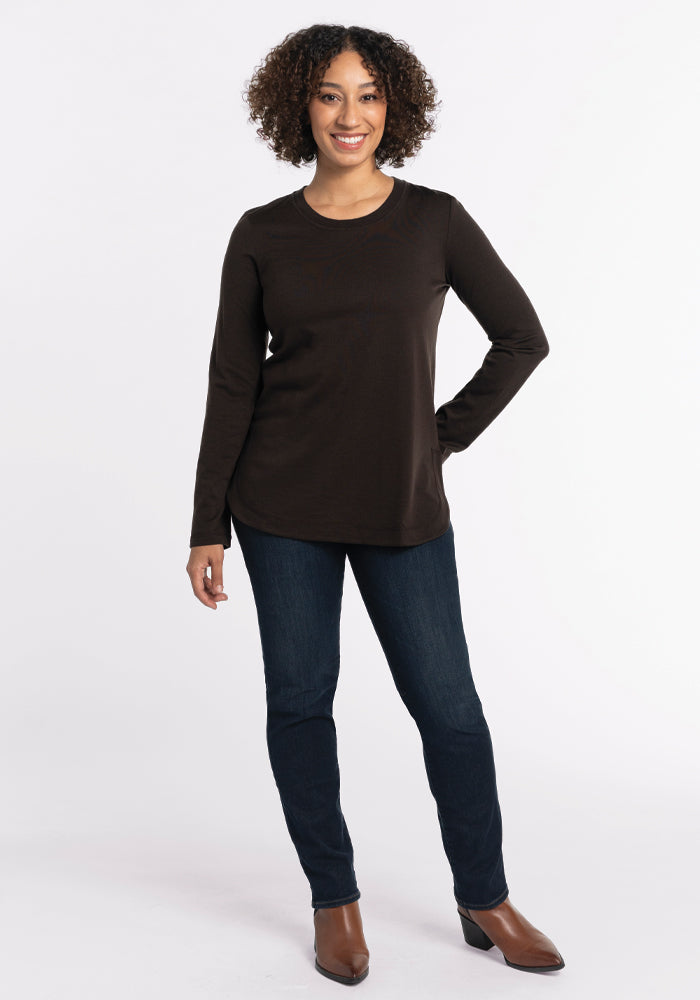 A person with curly hair smiles while wearing the Woolx Hazel Tunic in French Roast, along with dark jeans and brown shoes. They pose with a hand on their hip against a light background.