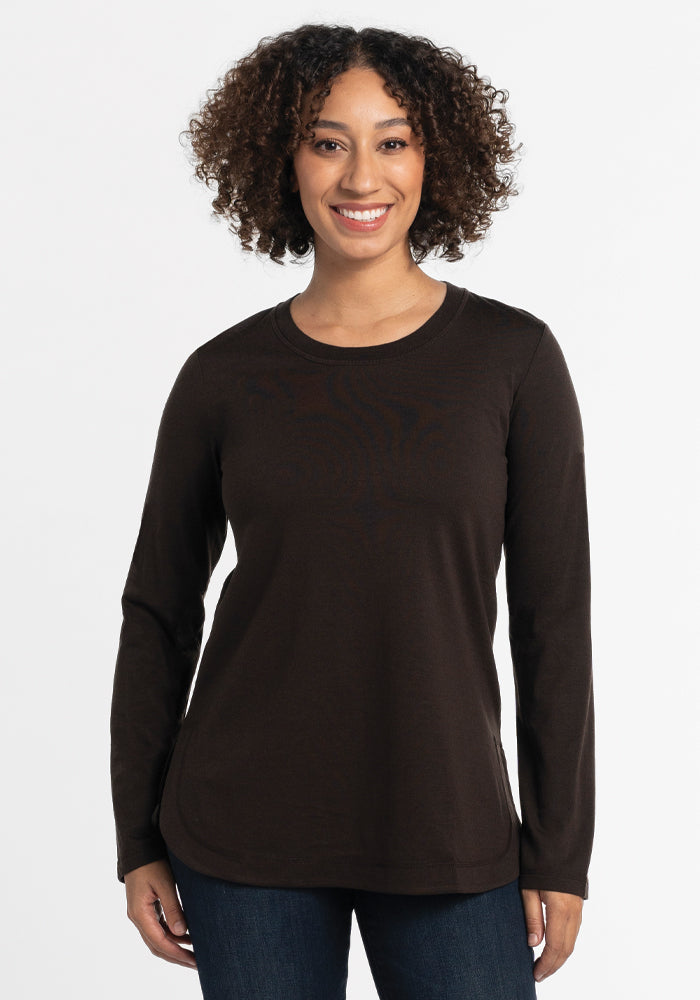 A person with curly hair smiles warmly, wearing the stylish Hazel Tunic - French Roast by Woolx, crafted from lightweight merino wool. Paired with jeans against a plain white background, they exude effortless style.