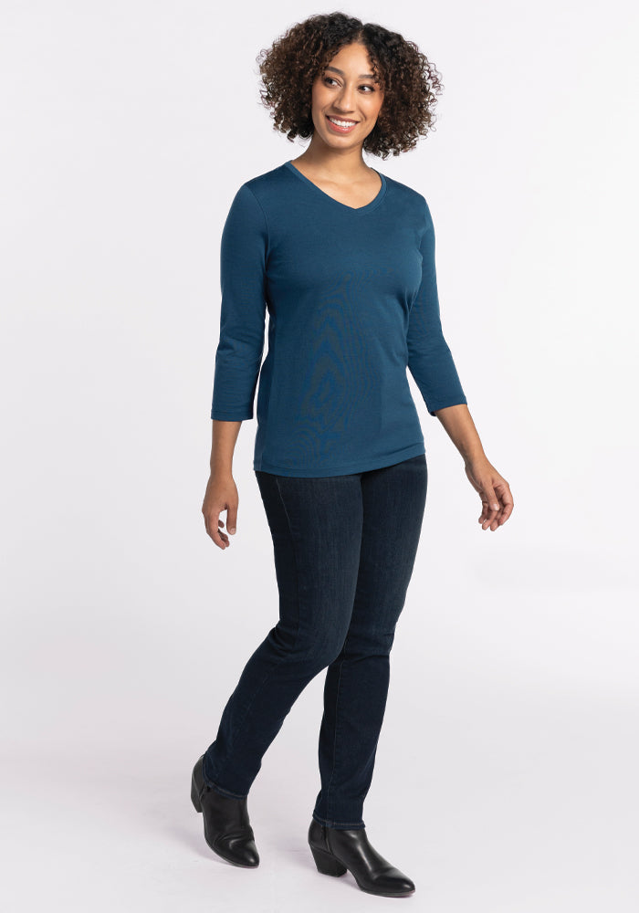 A person with curly hair walks confidently, showcasing the Woolx Elena V Neck in Real Teal, gracefully paired with dark jeans and black ankle boots, against a plain white background.