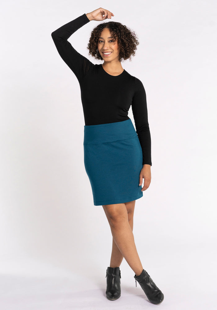 A person with curly hair stands confidently, wearing a black long-sleeve top and the Woolx Monroe Knee Length Skirt in Real Teal. They pose with one arm raised, smiling at the camera, completing their outfit with black ankle boots. Perfect for any travel wardrobe, the background is plain white.