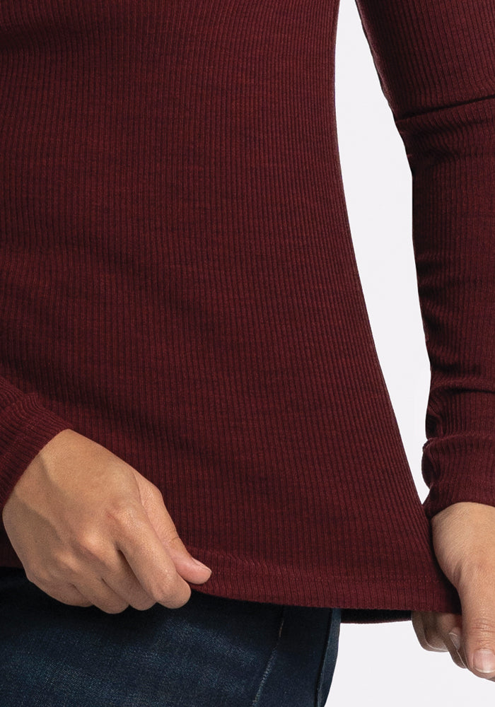 A person is wearing the Hadley Ribbed Crew in Cranberry Melange by Woolx, paired with dark jeans, and is gently pulling at the hem of the top with their right hand. The emphasis is on the texture and rich color of this travel wardrobe staple against a simple background.