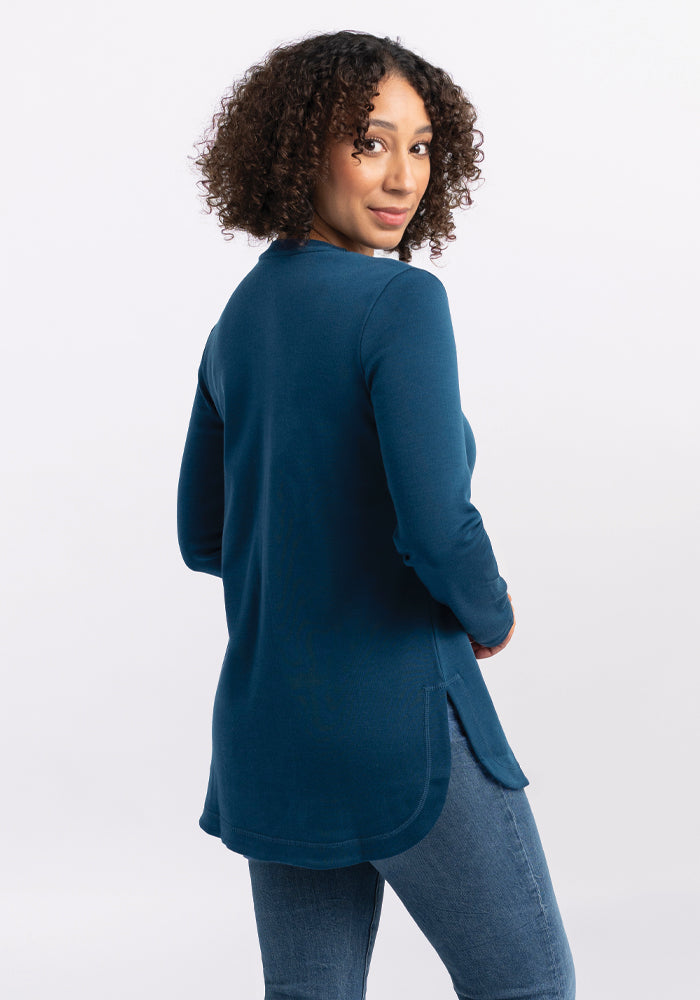 A person with curly hair, wearing the Woolx Hazel Tunic in Real Teal, a long-sleeve top made from lightweight fabric, and jeans, is turned slightly to the side, glancing back at the camera with a slight smile. The background is plain white.