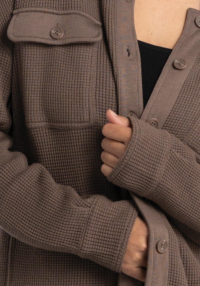 A close-up captures a person wearing the Woolx Oaklynn Waffle Shacket in Latte, draped over a black top. With its cozy neutral tone, this shacket is equipped with a buttoned chest pocket. The person's hand casually adjusts one side over the other.