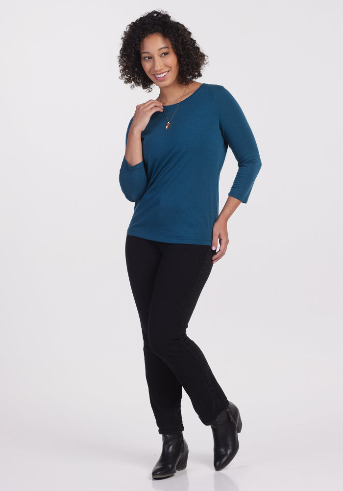 A person with curly hair, dressed in a lightweight Woolx Jenny 3/4 Sleeve Crew Neck top in Real Teal, black pants, and black ankle boots, poses with one hand on their chin and a slight smile. The background is plain white, lending a clean and simple look.