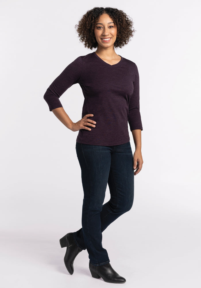 A person with curly hair stands confidently, wearing a relaxed fit Elena V Neck in Deep Plum from Woolx, jeans, and black ankle boots against a plain white background.