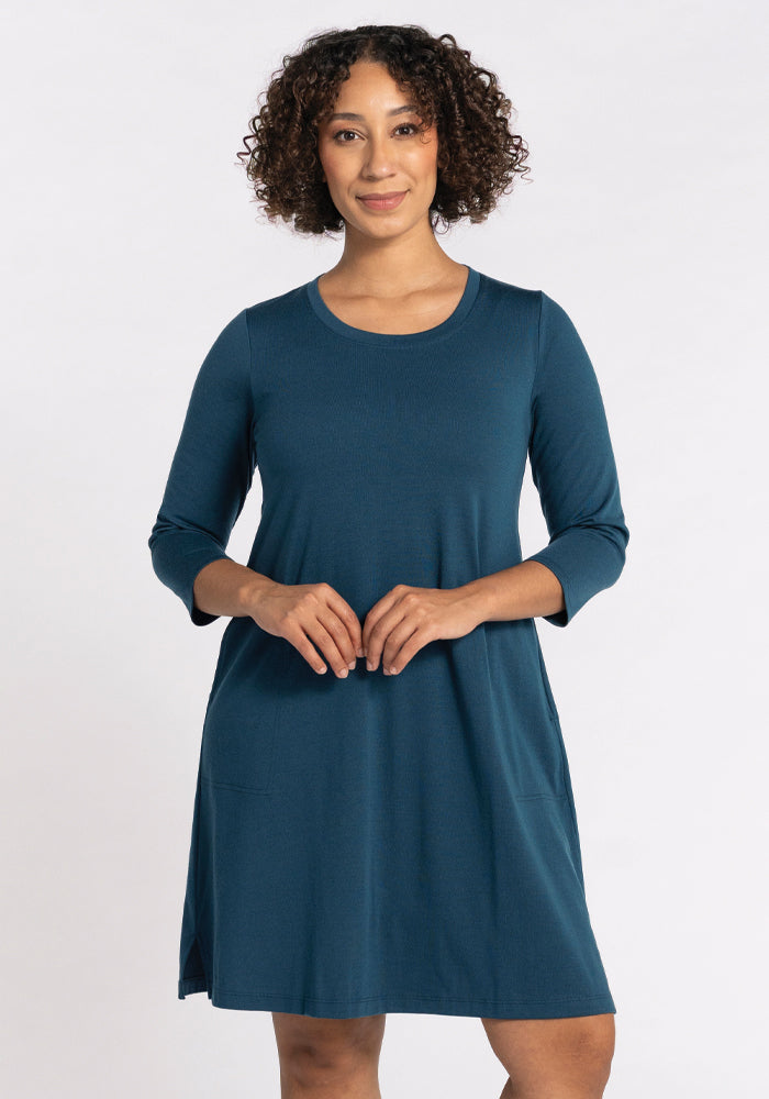 A person with curly hair is wearing the Rissa A Line Dress in Real Teal by Woolx, crafted from merino wool and showcasing three-quarter sleeves. They are standing against a plain white background, smiling, with their hands together at waist level. 