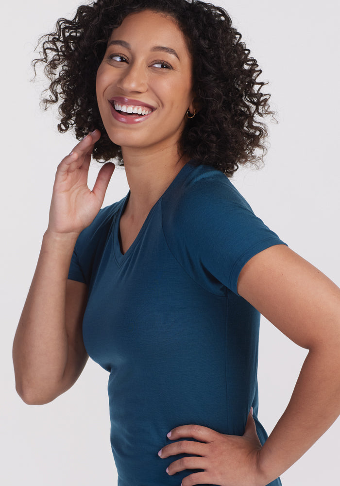 A woman with curly hair smiles while looking off to the side, dressed in the Woolx Mia V Neck shirt in Real Teal. She rests one hand near her neck and the other on her hip, all against a plain light background.