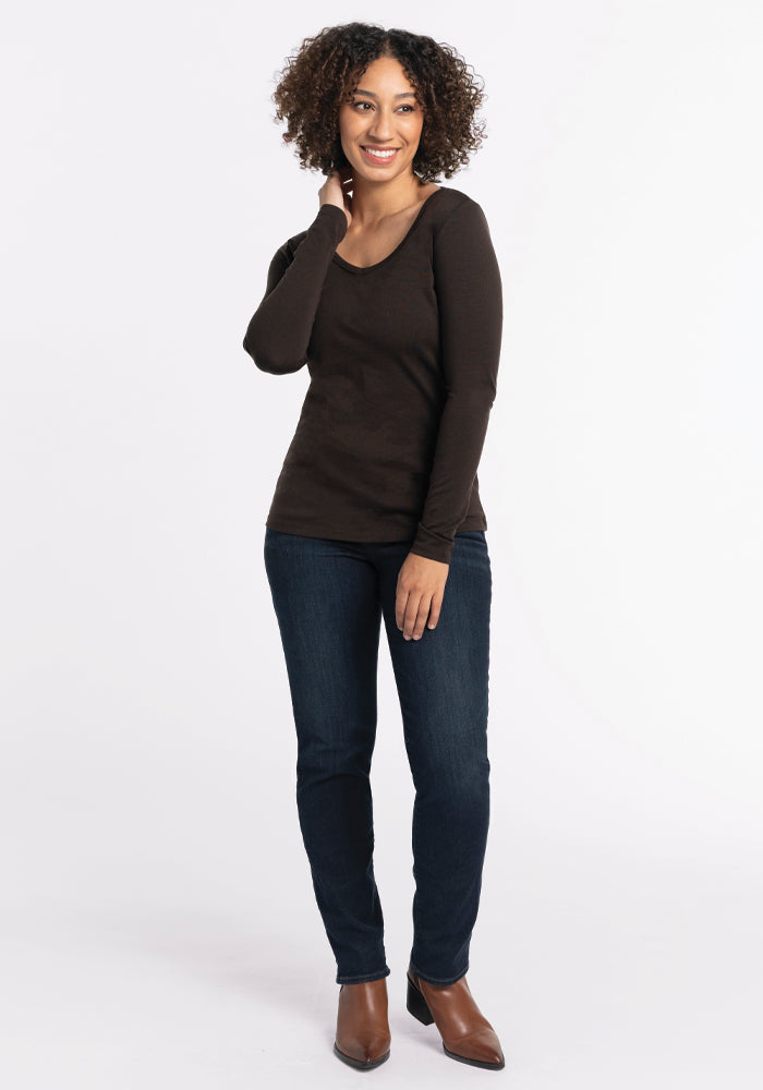 A person with curly hair is smiling and standing in a relaxed pose. They are wearing the Woolx Layla V Neck Top in French Roast, blue jeans, and brown boots against a plain white background.