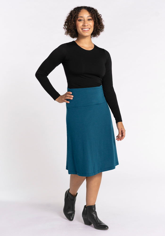 A person with curly hair wearing a black long-sleeve top, the Marilyn Flowy Midi Skirt in Real Teal from Woolx made of Merino wool, and black ankle boots poses confidently against a plain white background.