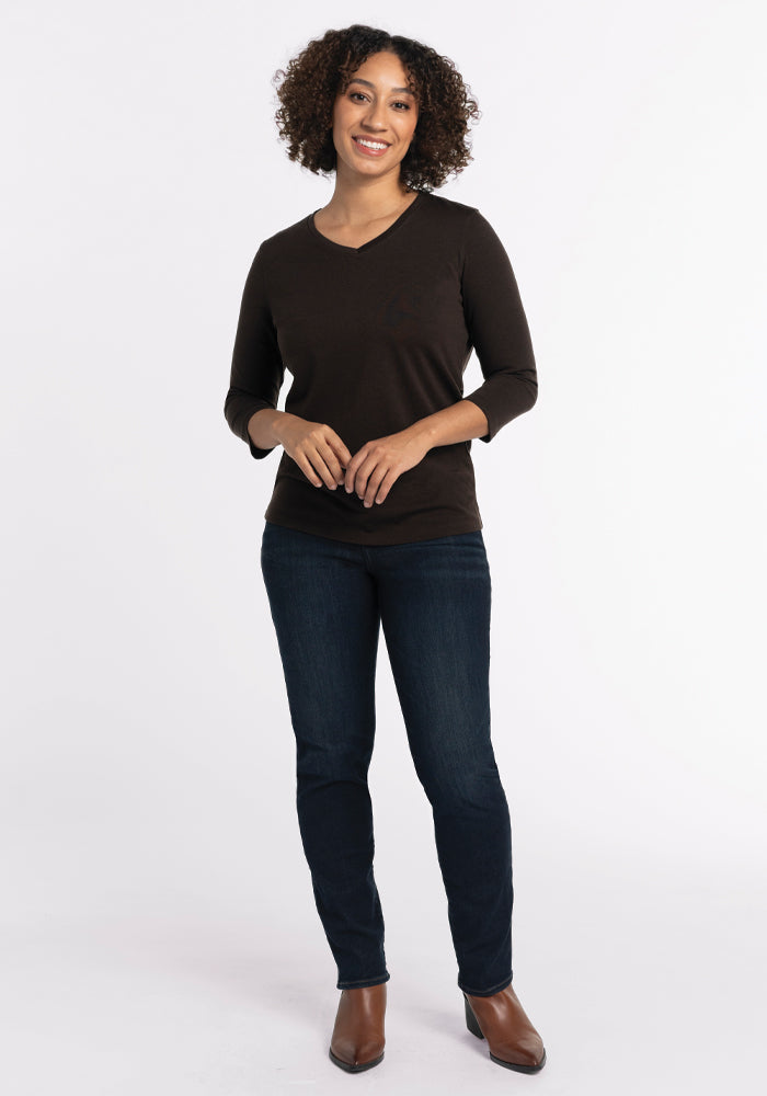 A person with curly hair is smiling while standing against a plain white background. They are wearing a relaxed fit, long-sleeve French Roast Elena V Neck top by Woolx, dark jeans, and brown shoes. Their hands are clasped in front of them.