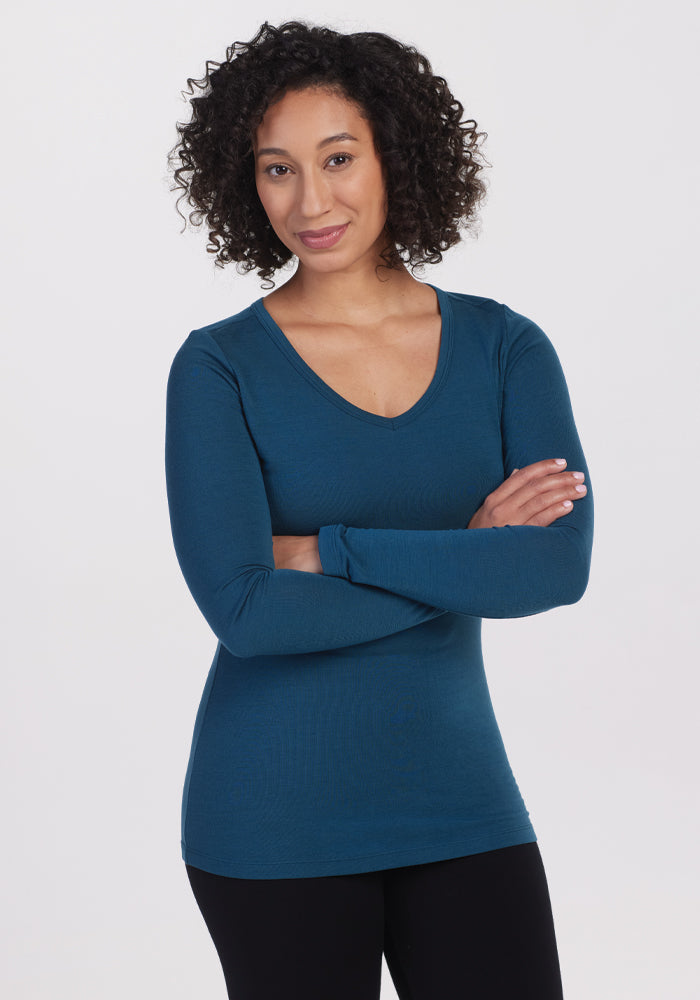 A person with curly hair is smiling, wearing the Woolx Layla V Neck Top in Real Teal and black pants. They have their arms crossed and are standing against a plain white background.