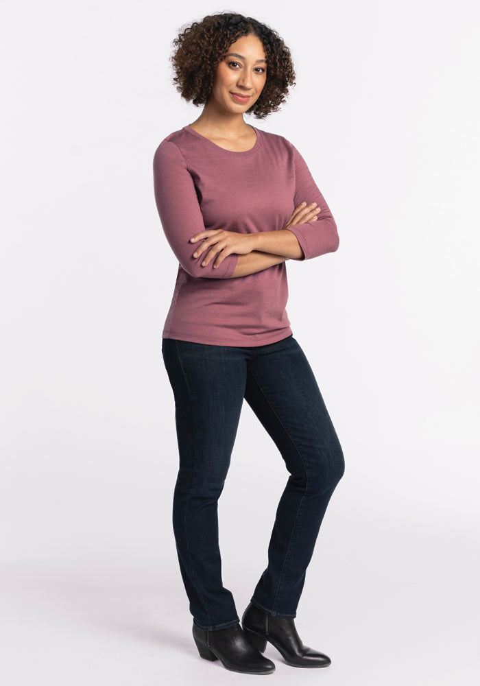 A person with curly hair stands confidently with arms crossed, wearing a Wild Ginger Jenny 3/4 Sleeve Crew Neck by Woolx, paired with dark jeans and black shoes. The background is plain white.