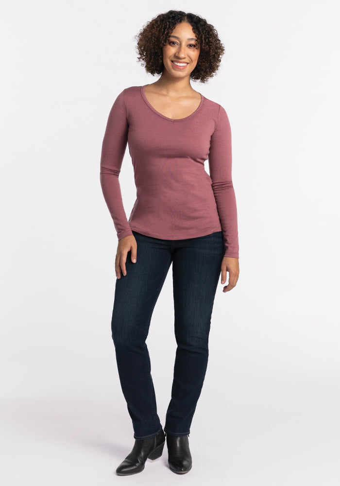 A person with curly hair stands smiling, dressed in a cozy Woolx Layla V Neck Top in Wild Ginger, dark jeans, and sleek black ankle boots. The clean white backdrop highlights their casual yet stylish ensemble.