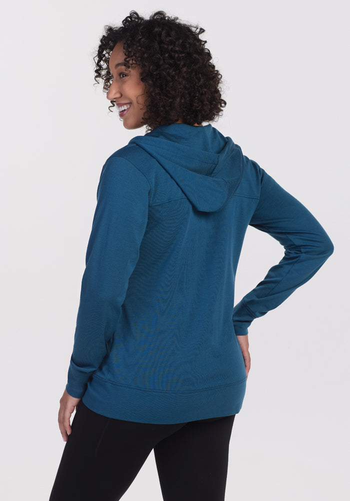 A person with curly hair is smiling while wearing the Woolx Ryann Hoodie in Real Teal, showcasing its lightweight, athletic fit from the back. They are standing against a plain white background.