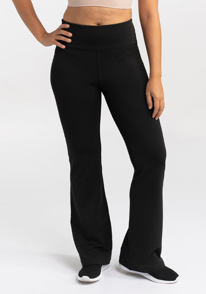 A person wearing Woolx's Marissa Pants - Black and black sneakers stands with one hand on their hip. The high-waisted pants are complemented by a cropped top that reveals their midriff, set against a plain, light background.