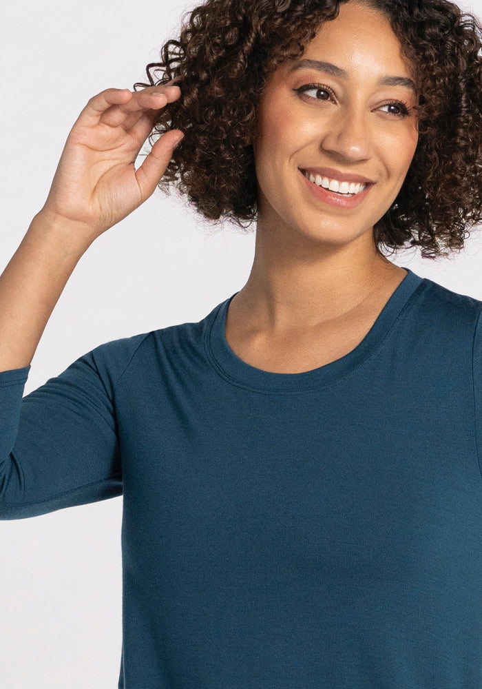 A person with curly hair is smiling and wearing the travel-ready Rissa A Line Dress in real teal from Woolx. They are looking slightly to the side against a plain, light background.