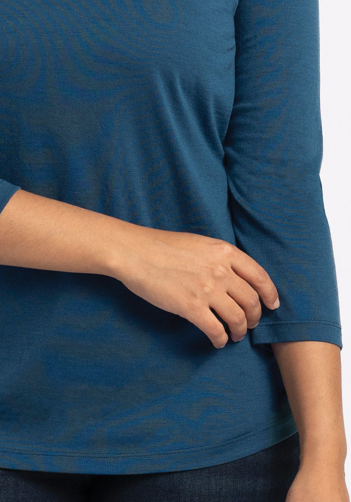 Someone is wearing a Real Teal Elena V Neck from Woolx, shown from the shoulders to the hips. Their left hand gently rests on their right arm, emphasizing the relaxed fit and texture of this shirt made from soft Merino wool.