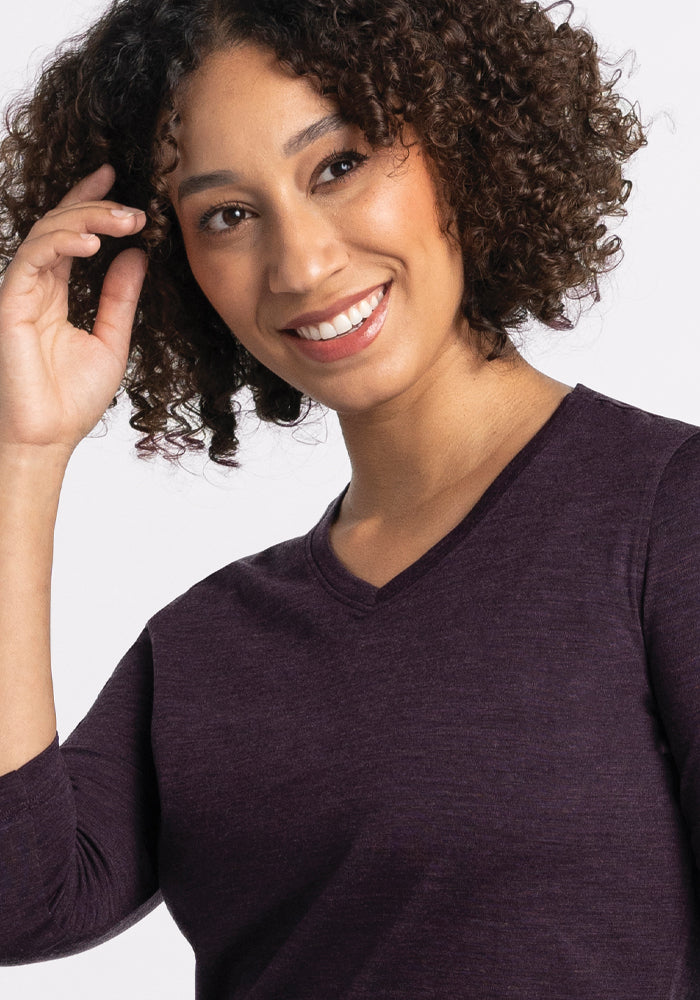 A person with curly hair smiles warmly, wearing a relaxed-fit Elena V Neck from Woolx in deep plum. They touch their hair with one hand against a plain light background.