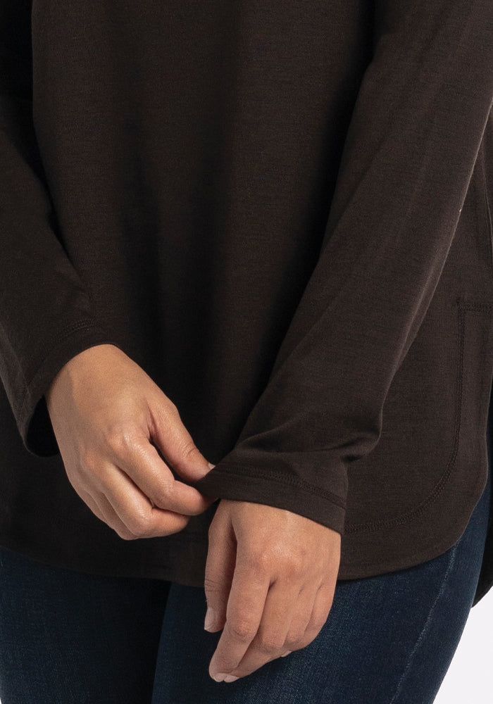 Dressed in the lightweight Woolx Hazel Tunic - French Roast and a pair of jeans, someone is adjusting the cuff of their sleeve with one hand while holding the fabric with the other. The background is plain and light-colored.