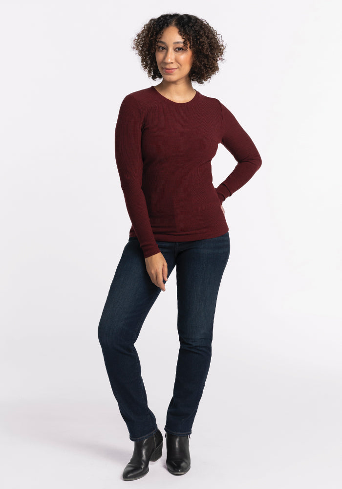 A person with curly hair stands against a white background, wearing the Hadley Ribbed Crew in Cranberry Melange by Woolx, paired with dark jeans and black ankle boots. They smile warmly, extending one hand forward while casually resting their weight on one leg. It's the perfect addition to any travel wardrobe.