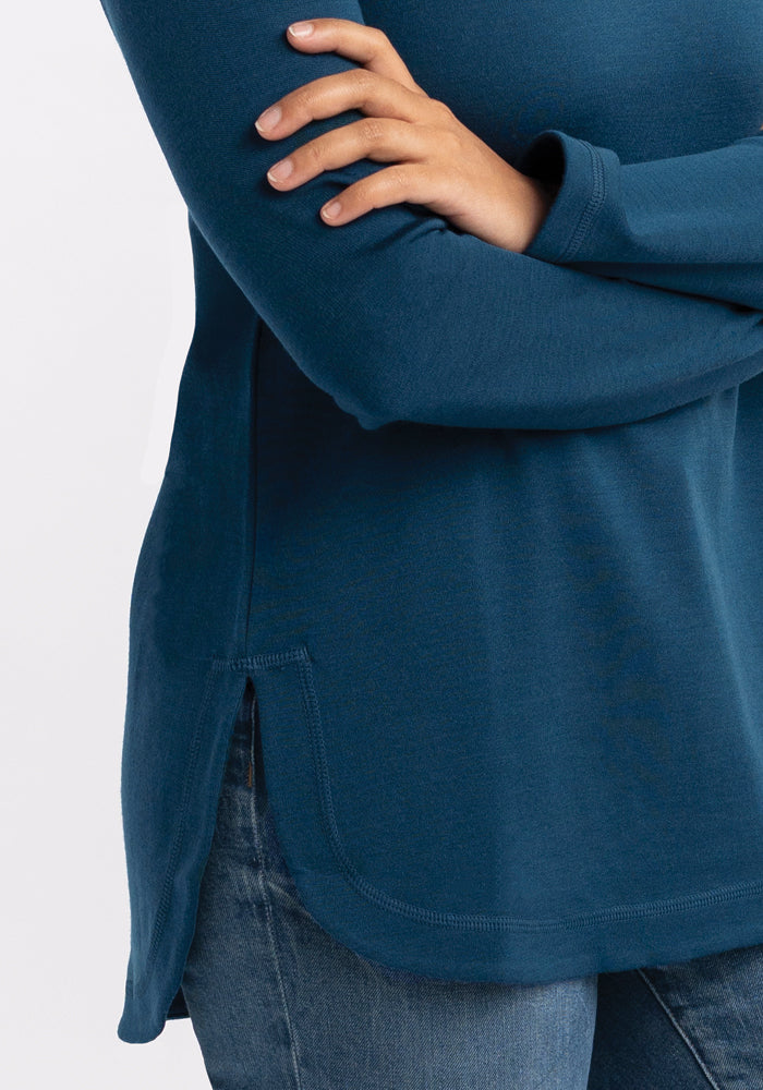 The individual is seen wearing a Woolx Hazel Tunic in Real Teal, long-sleeved with arms crossed, depicted from the shoulder to the waist. This tunic showcases a rounded hem and side slit, made from lightweight fabric. They have paired it with blue jeans and are posed against a simple white background.