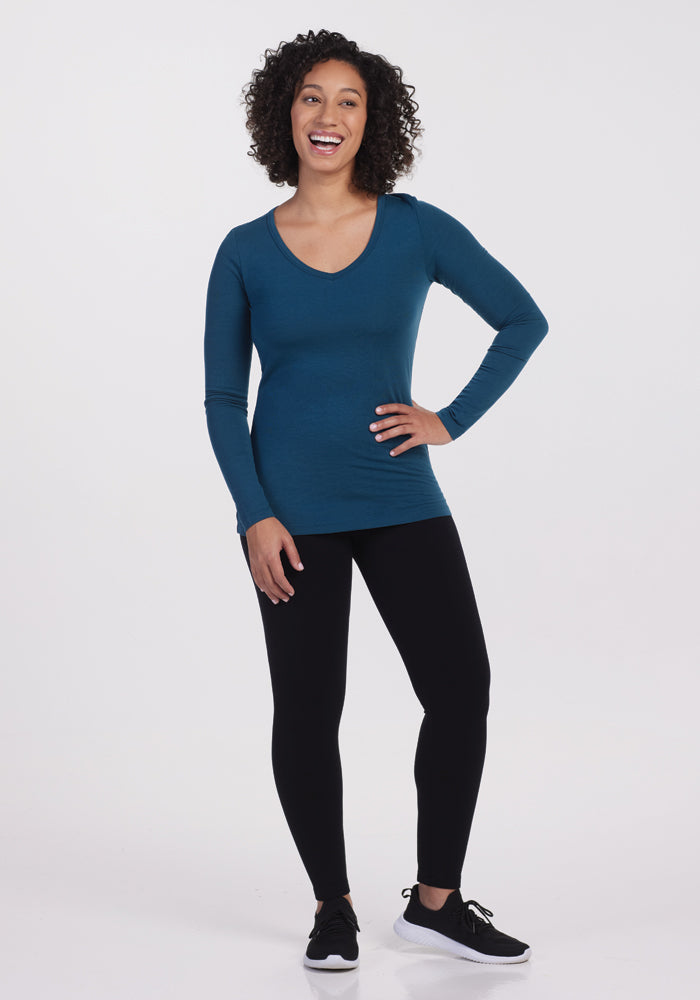 A person with curly hair is standing and smiling, dressed in a Woolx Layla V Neck Top in Real Teal, black leggings, and black sneakers against a plain white background.