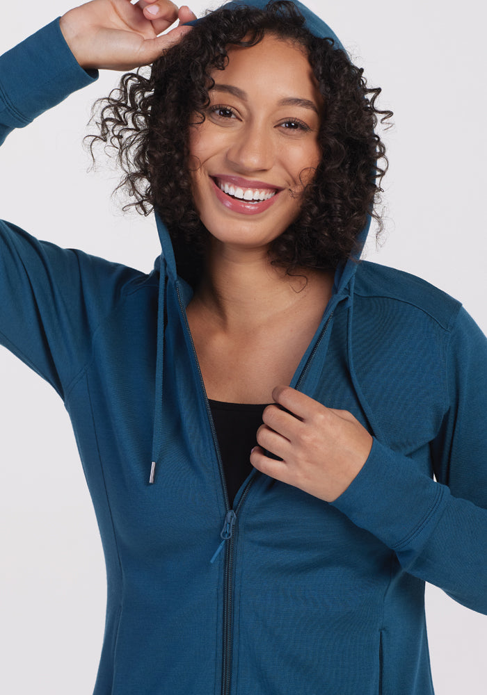 A person beams as they wear the Woolx Ryann Hoodie in Real Teal, gently pulling the hood over their curly hair. With its athletic fit, this Merino wool hoodie pops against the plain white backdrop.