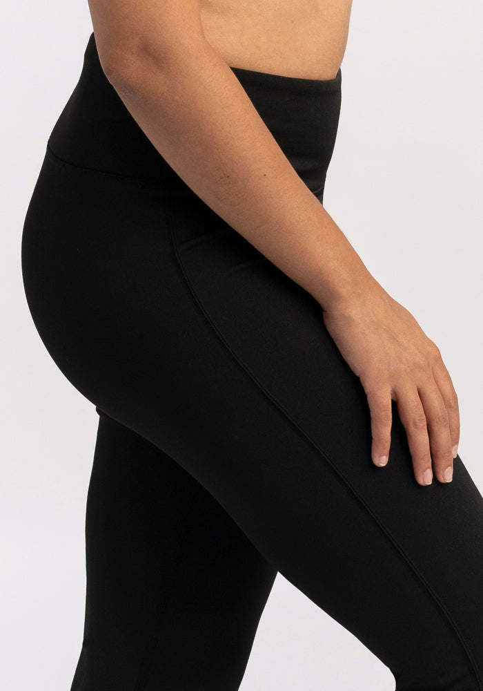 A person wearing Woolx's Marissa Pants in black is shown from the side, with their right arm resting at their side. The background is plain and light-colored.