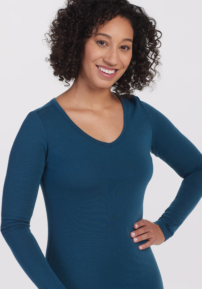 A smiling person with curly hair is posing with one hand on their hip against a plain white background while wearing the Woolx Layla V Neck Top in Real Teal.