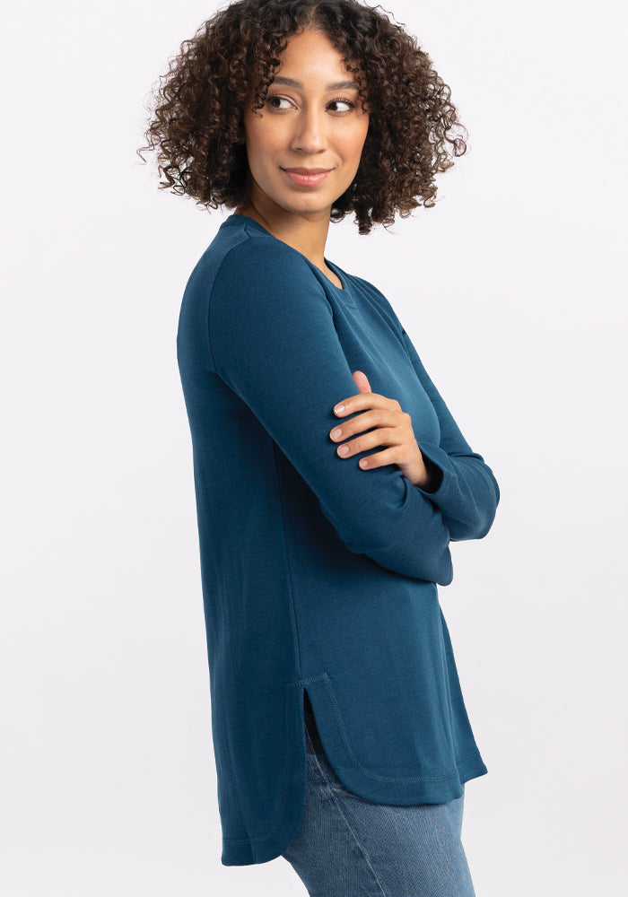 A person with curly hair is smiling over their shoulder, dressed in a real teal Hazel Tunic by Woolx, made from lightweight fabric, paired with jeans, as they stand against a plain white background.